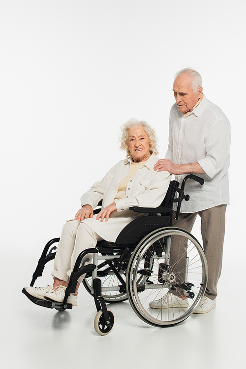 elderly man holding hand on shoulder of wife in wheelchair isolated on white