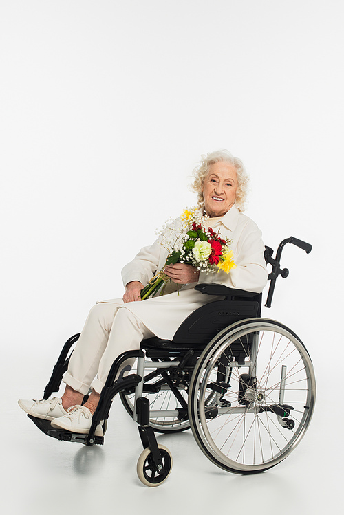 smiling elderly woman in casual clothes sitting in wheelchair holding bouquet of flowers on white