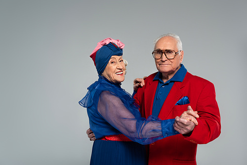 smiling elderly couple in blue dress and red blazer dancing isolated on grey