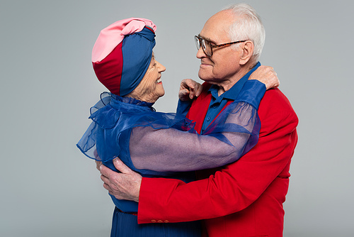 smiling elderly couple in blue dress and red blazer hugging isolated on grey