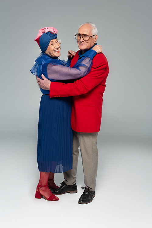 smiling elderly couple in blue dress and red blazer hugging on grey