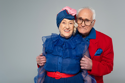 smiling elderly man in red blazer hugging wife in blue dress and turban isolated on grey