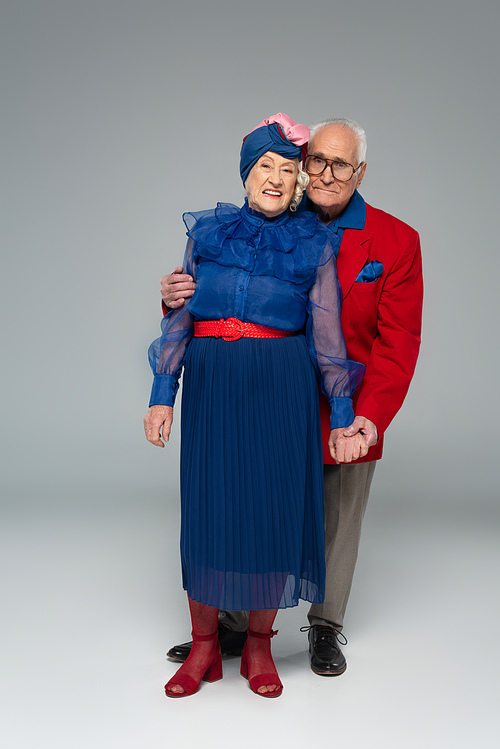 smiling elderly couple in blue dress and red blazer hugging and holding hands on grey