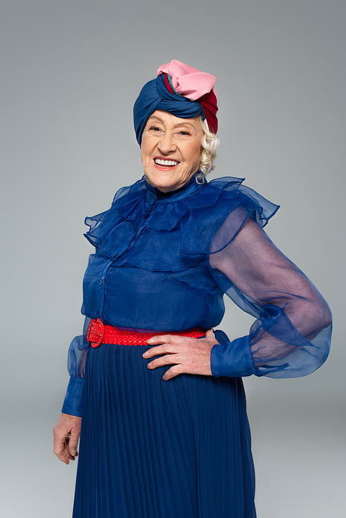 smiling elderly woman in blue dress and turban posing with hand on hip isolated on grey
