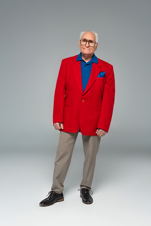 full length view of serious elderly man in glasses, red blazer and blue shirt standing on grey
