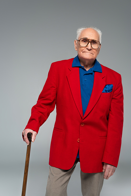 serious elderly man in red blazer and glasses standing with walking stick isolated on grey