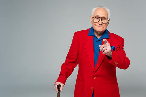 elderly man in red blazer standing with walking stick and pointing with finger to camera isolated on grey