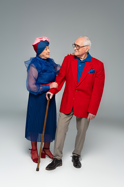 smiling elderly woman in blue dress hugging husband in red blazer with walking stick on grey