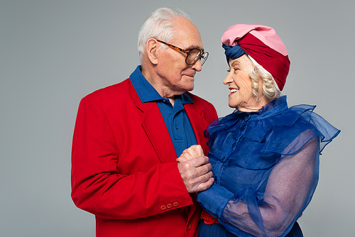 smiling elderly man in red blazer holding hands with wife in blue dress and turban isolated on grey