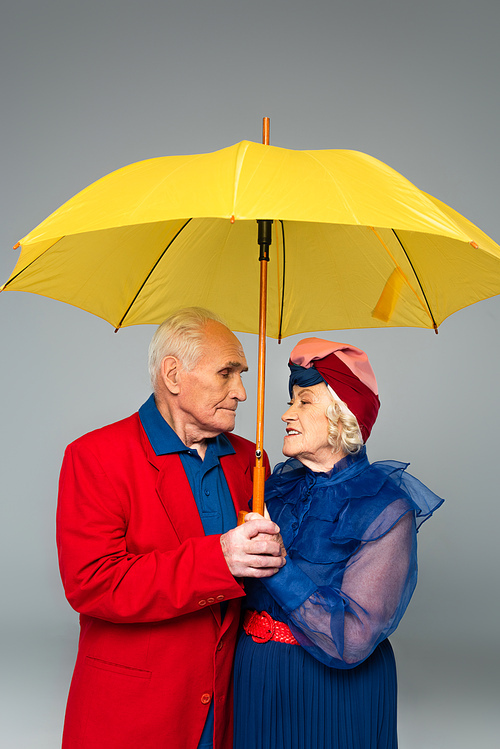 elderly man in red blazer and woman in blue dress and turban holding yellow umbrella isolated on grey
