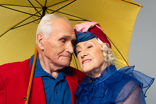 elderly man with yellow umbrella hugging wife in blue dress and turban isolated on grey
