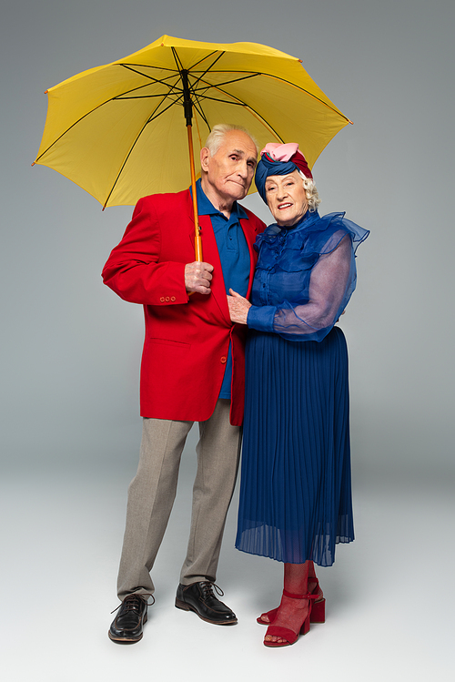 elderly man in red blazer with yellow umbrella hugging wife in blue dress and turban on grey