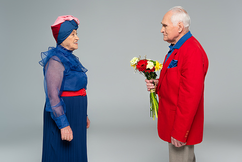 elderly man in red blazer holding bouquet of flowers near wife in blue dress and turban on grey