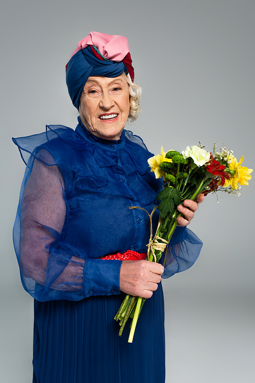 smiling elderly woman in blue dress and turban holding bouquet of flowers isolated on grey