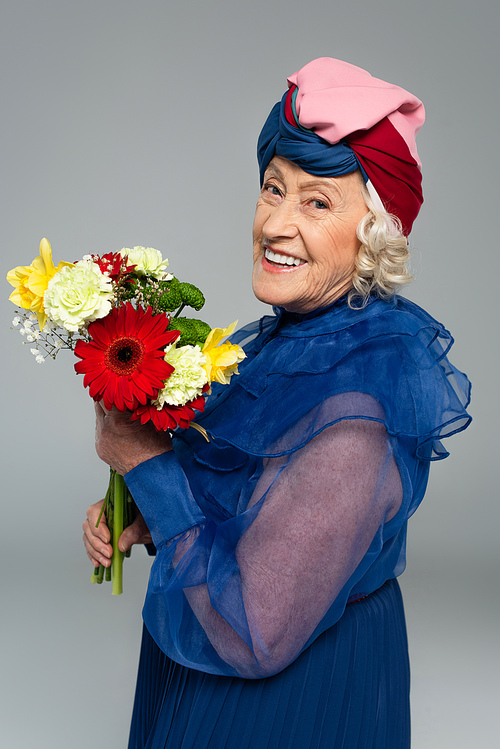 positive elderly woman in blue dress and turban holding bouquet of flowers isolated on grey