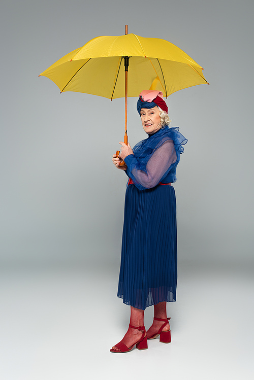 smiling elderly woman in blue dress and turban holding yellow umbrella on grey