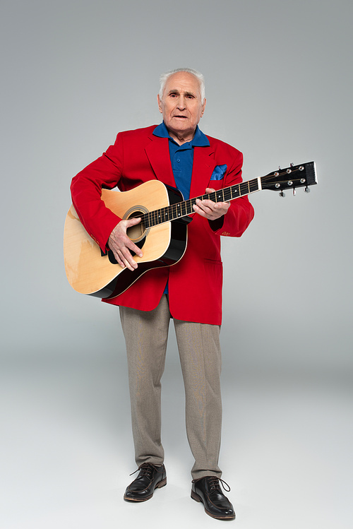 elderly man in red blazer and brown trousers holding acoustic guitar on grey