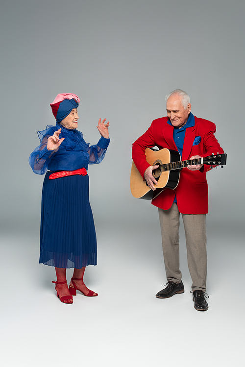 positive elderly man in red blazer playing acoustic guitar near dancing woman in blue dress and turban on grey