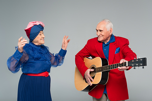 smiling elderly man in red blazer playing acoustic guitar near dancing woman in blue dress isolated on grey