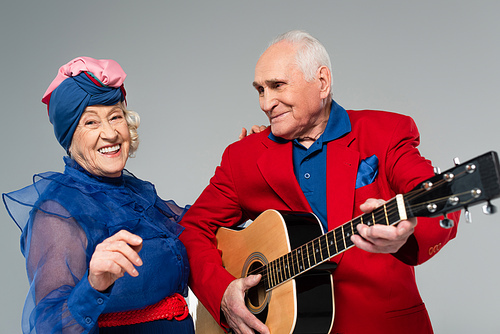 positive elderly man in red blazer playing acoustic guitar near dancing woman in blue dress and turban isolated on grey