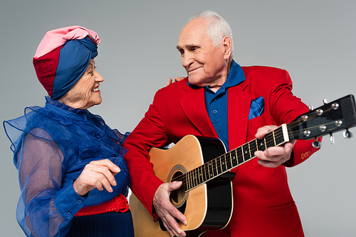 smiling elderly man in red blazer playing acoustic guitar near dancing woman in blue dress and turban isolated on grey