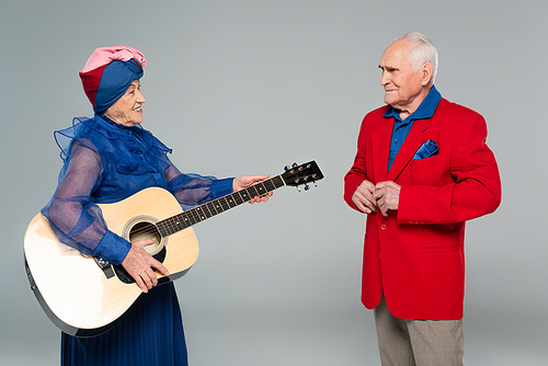 smiling elderly woman in blue dress and turban holding acoustic guitar near man in red blazer isolated on grey