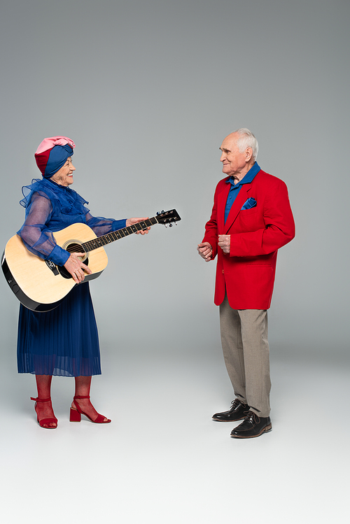 positive elderly couple in stylish clothes dancing with acoustic guitar on grey