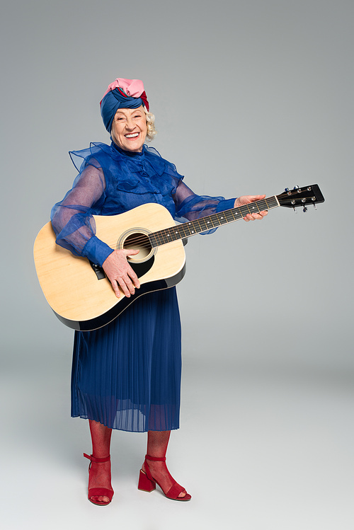 smiling elderly woman in blue dress and turban holding acoustic guitar on grey