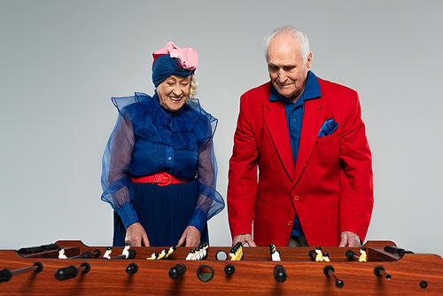 positive elderly couple in stylish clothes playing table football on grey