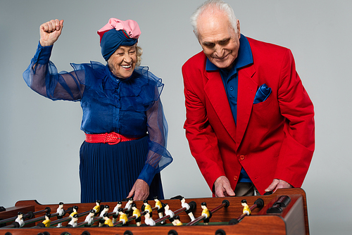 excited elderly couple in stylish clothes playing table football with win gesture on grey