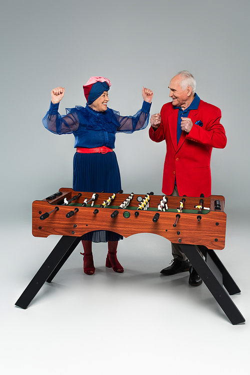 excited elderly couple in stylish clothes playing table football with win gestures on grey