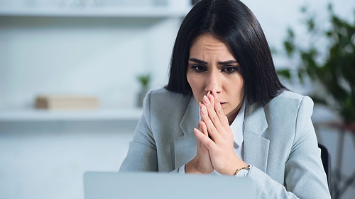 worried businesswoman with praying hands near blurred laptop in office