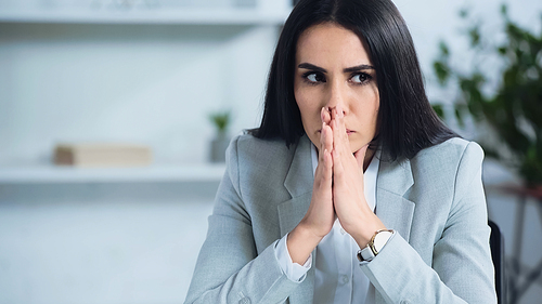 worried woman with praying hands near blurred laptop in office