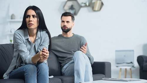 upset woman sitting with clenched hands near blurred and displeased boyfriend