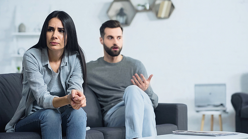 worried woman sitting with clenched hands near blurred and displeased boyfriend