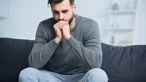 worried man in grey sweater sitting with clenched hands on couch