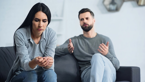 worried woman sitting and looking at hands near blurred and displeased boyfriend quarrelling in living room