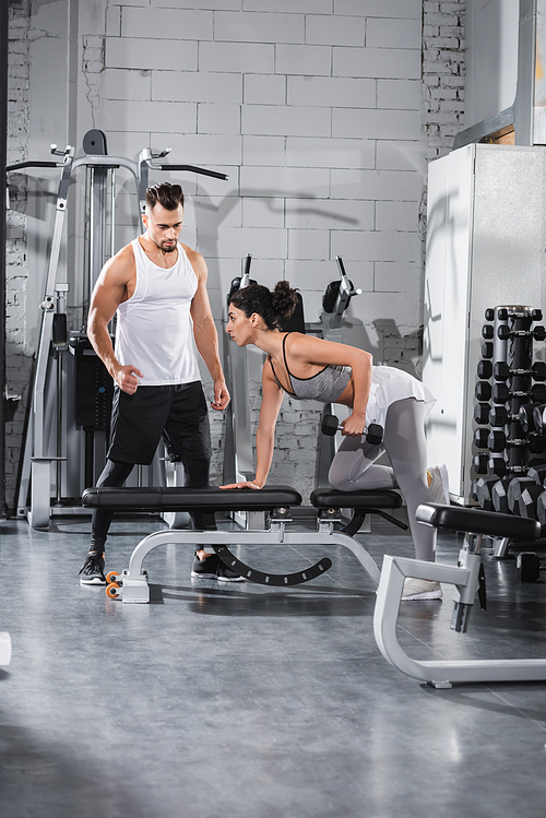 Middle east sportswoman working out with dumbbell on flat bench near trainer in gym