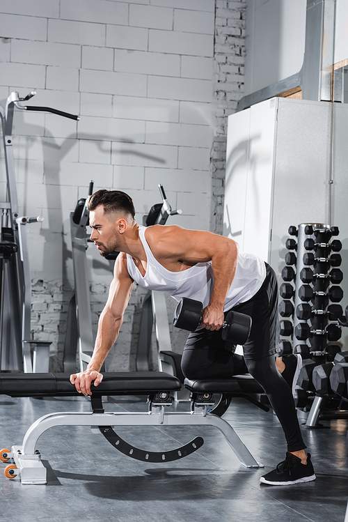 Side view of athletic sportsman training with dumbbell on flat bench in gym