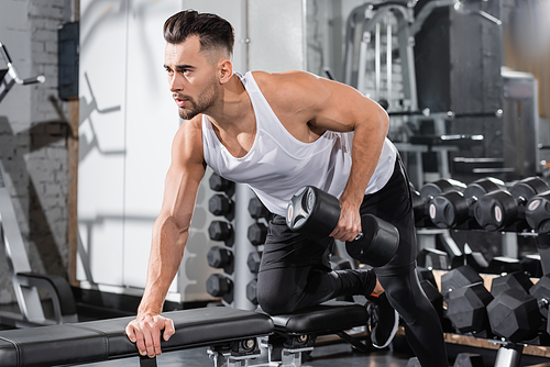 Young sportsman working out with dumbbell on flat bench in gym