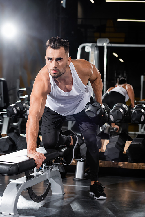 Athletic sportsman working out with dumbbell near flat bench in gym