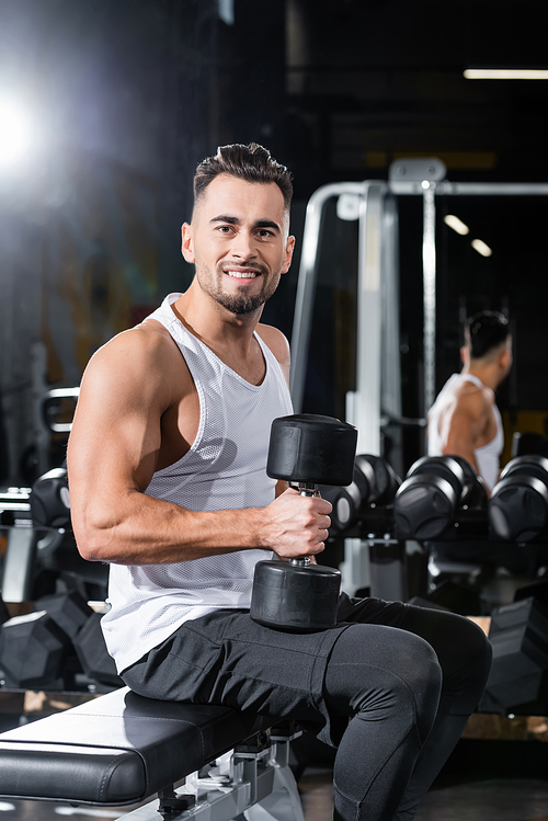 Smiling sportsman holding dumbbell while sitting on flat bench in gym