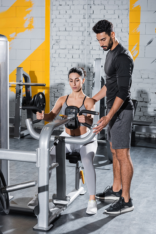 Arabian trainer standing near middle east sportswoman training on ab crunch machine in gym