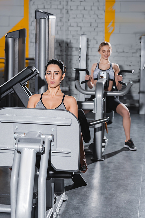 Young middle east sportswoman working out on leg press machine near blurred friend in gym