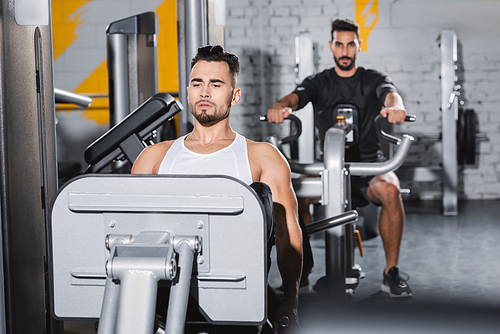 Sportsman training on leg press machine near blurred muslim friend in gym