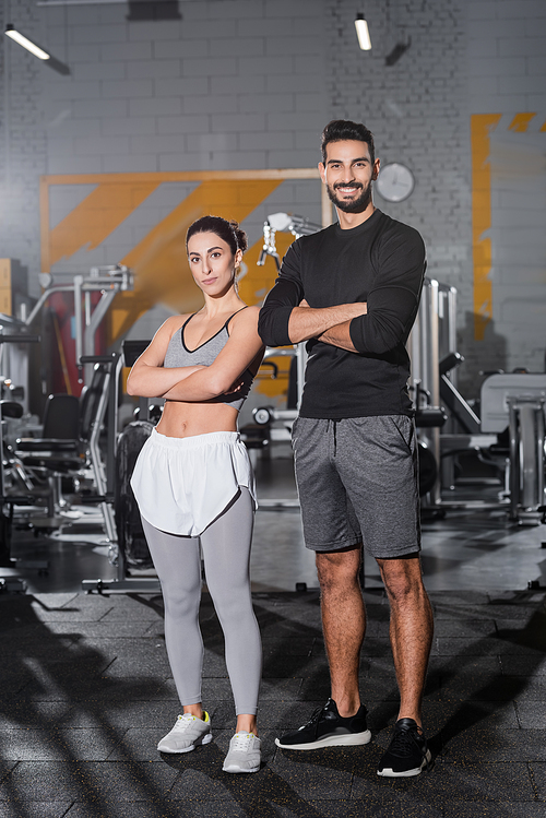 Positive muslim sportsman crossing arms near middle east sportswoman in gym