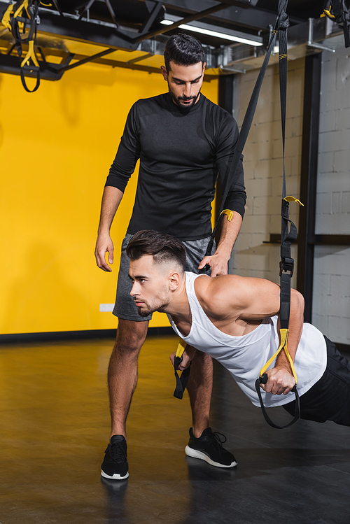 Arabian trainer standing near friend working out with suspension straps in gym