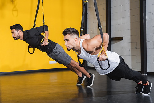 Young multiethnic sportsmen training with suspension straps in gym