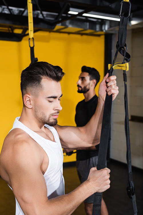 Young man holding suspension straps near blurred arabian friend in gym