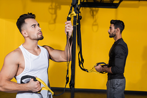 Young man holding suspension straps near blurred arabian friend in gym